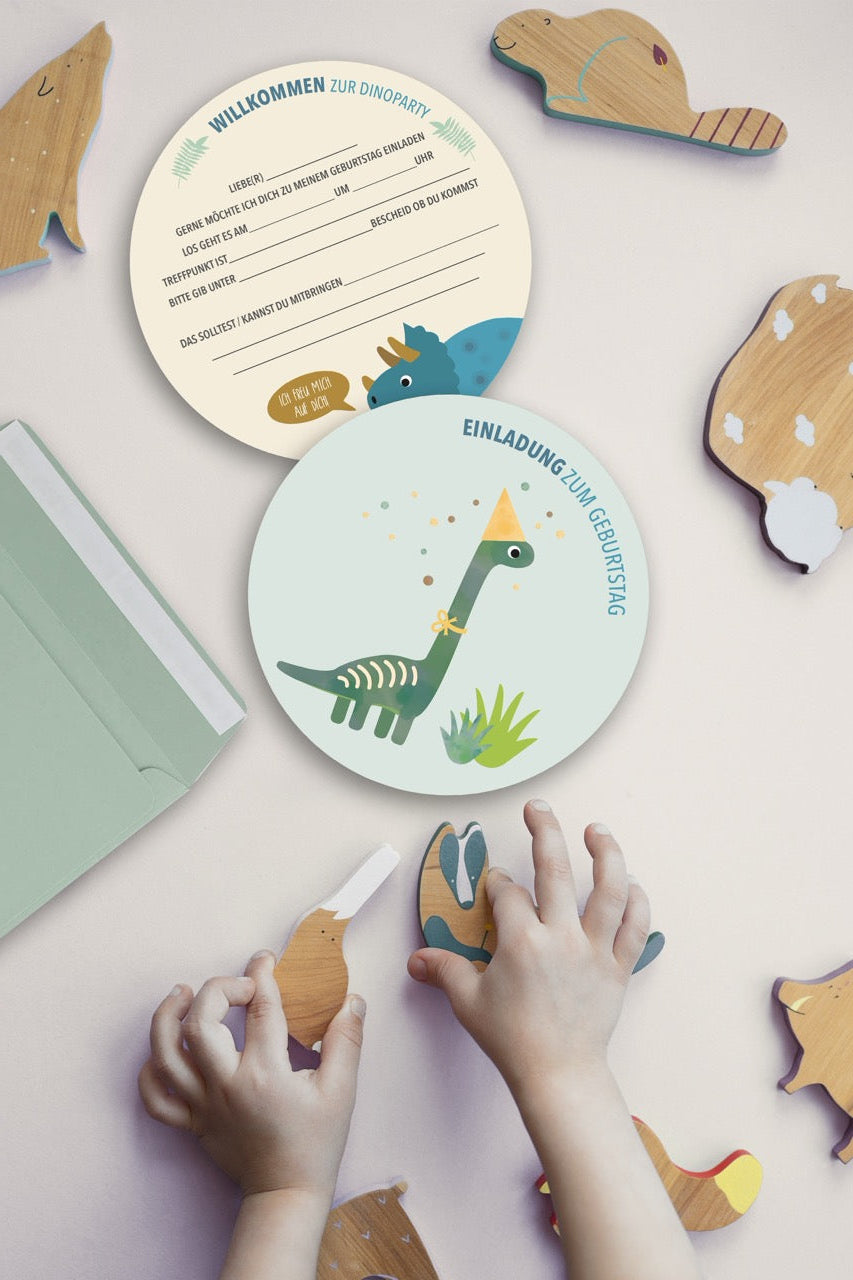 Zwei runde Einladungskarten zu einem Kindergeburtstag mit Dinosaurier-Motiv liegen auf einem Tisch. Eine Karte zeigt einen bunten Dinosaurier, die andere enthält Textfelder für Einladungshinweise. Neben den Karten greifen Kinderhände nach Dinosaurier-Puzzleteilen aus Holz. Eine grüne Umschlag liegt unter den Karten.