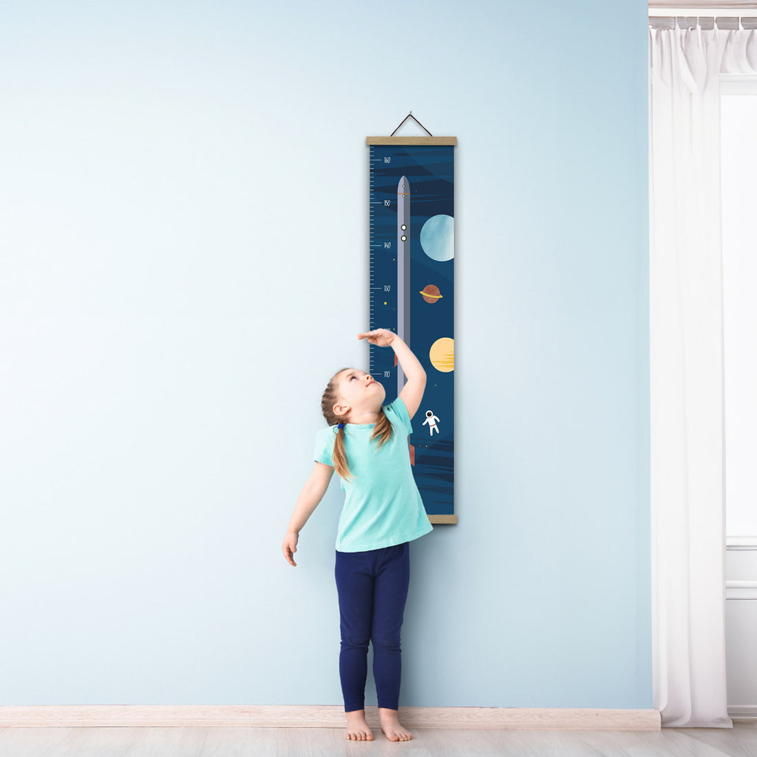 Ein Mädchen in blauer Kleidung steht barfuß auf einem Holzboden. Sie misst ihre Größe an einem mit einer Rakete illustrierten Messlatte an einer blauen Wand.