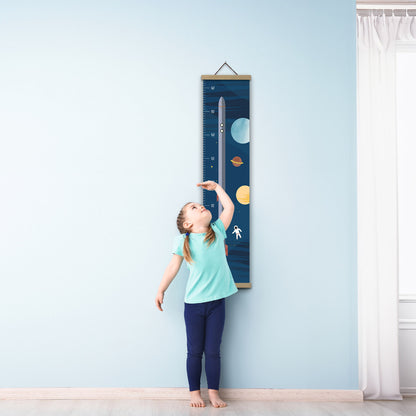 Ein Mädchen in blauer Kleidung steht barfuß auf einem Holzboden. Sie misst ihre Größe an einem mit einer Rakete illustrierten Messlatte an einer blauen Wand.