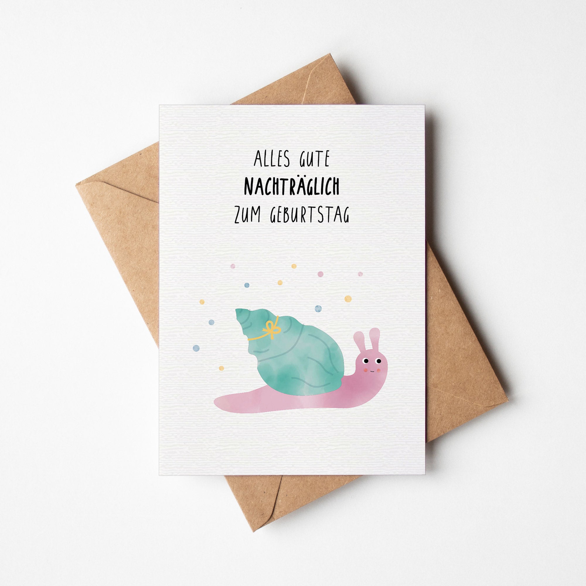 Nachträgliche Geburtstagskarte mit einer niedlichen Illustration einer Schnecke und dem Text &quot;Alles Gute nachträglich zum Geburtstag&quot;. Die Schnecke hat ein großes, grünes Schneckenhaus mit einer Schleife und ist von bunten Konfetti umgeben. Die Karte liegt auf einem braunen Umschlag.